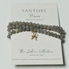 Gray beaded bracelet with a gold charm on a Santore branded card.